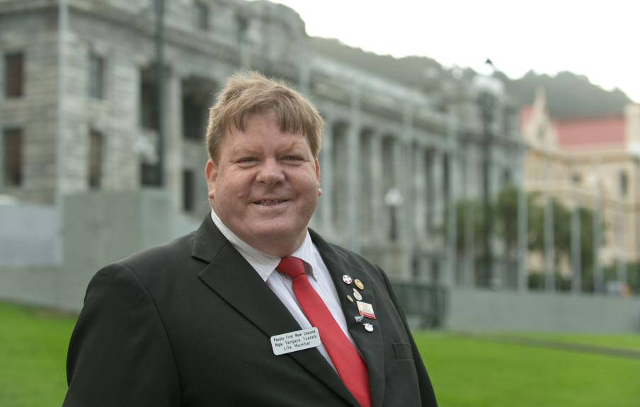 Robert Martin in a black suit with a red tie smiling at the camera and standing in front of the New Zealand Parliament building.