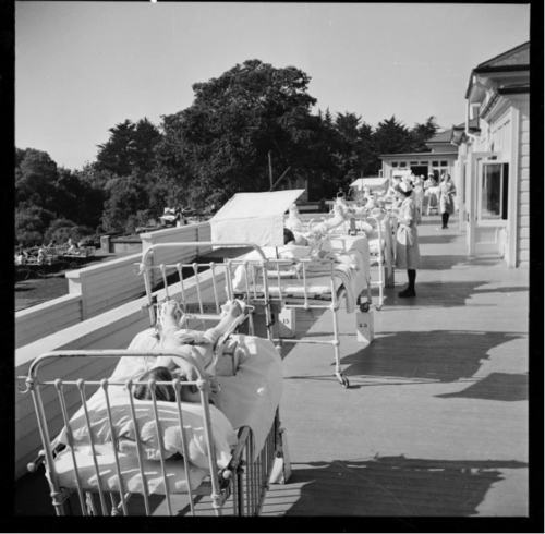 A black and white photo of children lying metal hospital beds outside on a terrace, attended by nurses